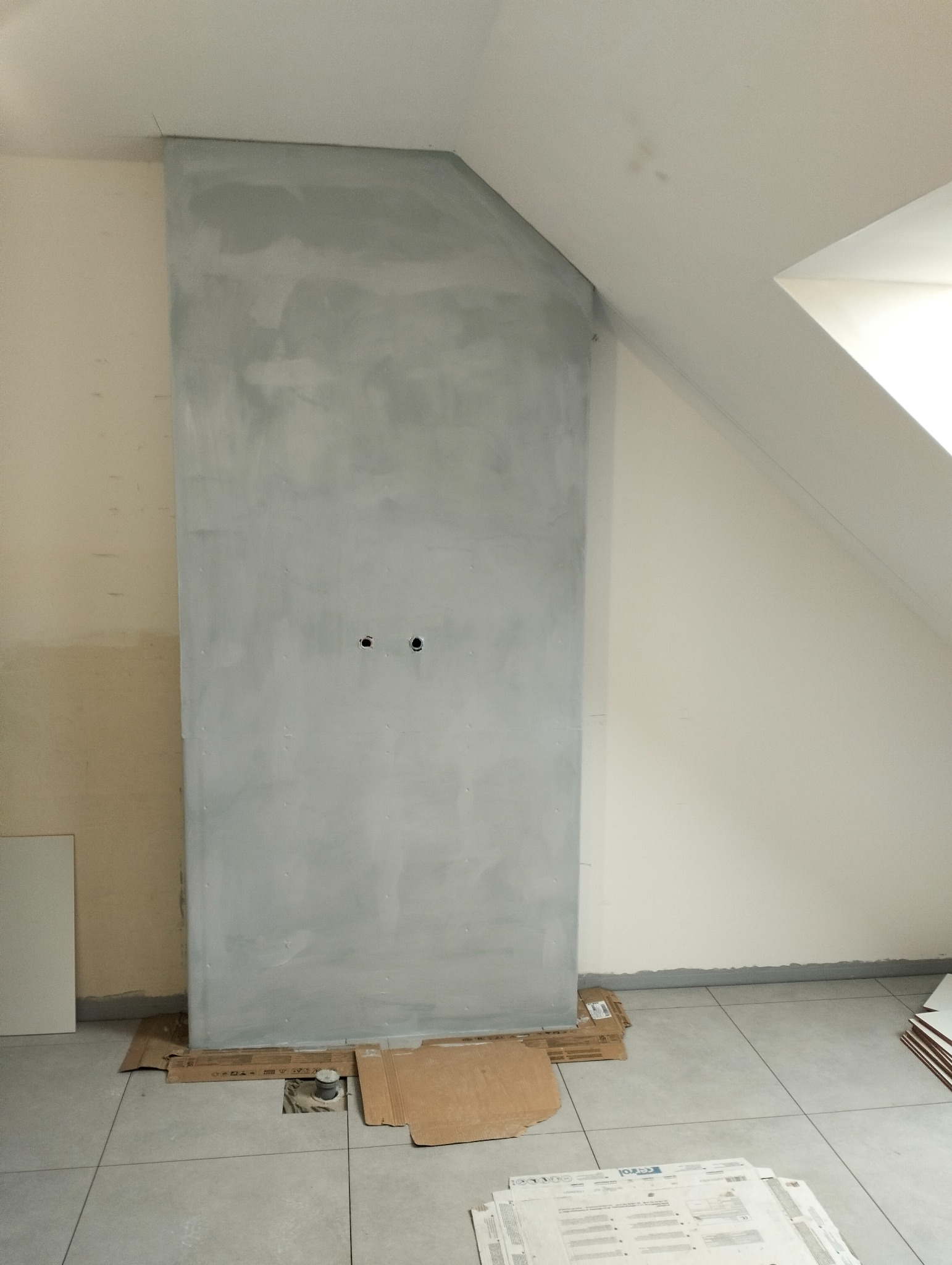 Ściana po szpachlowaniu w szarym kolorze, przygotowana pod malowanie, w pokoju na poddaszu. Widoczne otwory na gniazdka elektryczne i fragmenty płytek podłogowych.