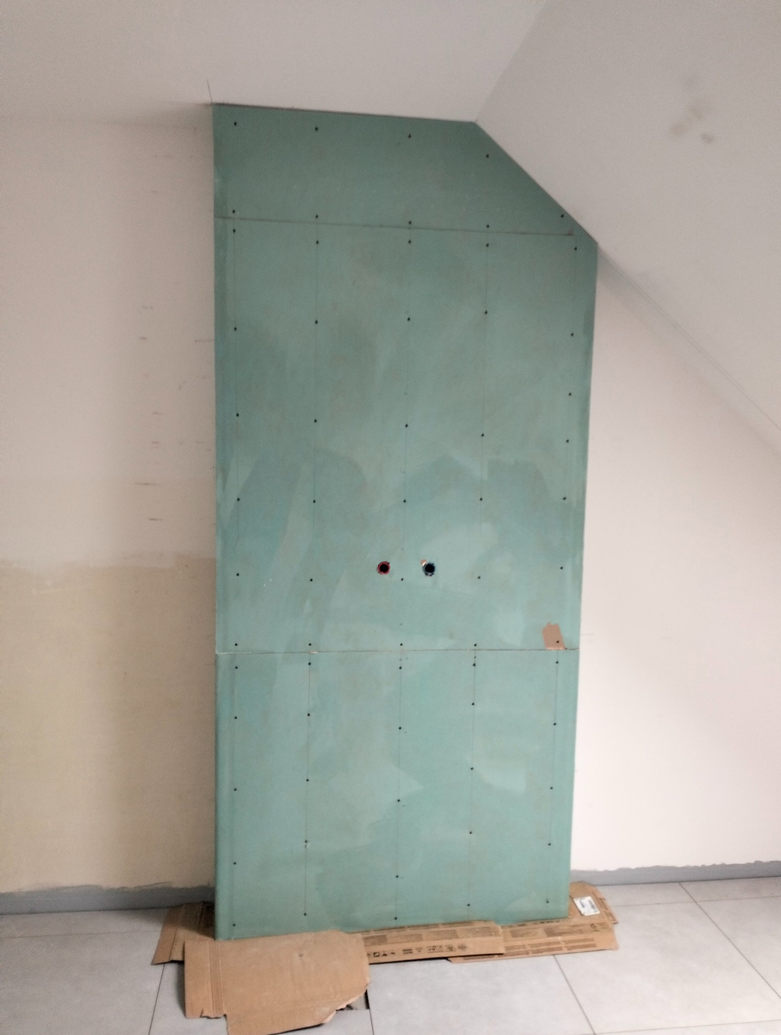 Ściana z płyty karton-gips w skosie poddasza z widocznymi otworami na instalacje, przymocowana do ściany, na podłodze kartony zabezpieczające przed uszkodzeniami.