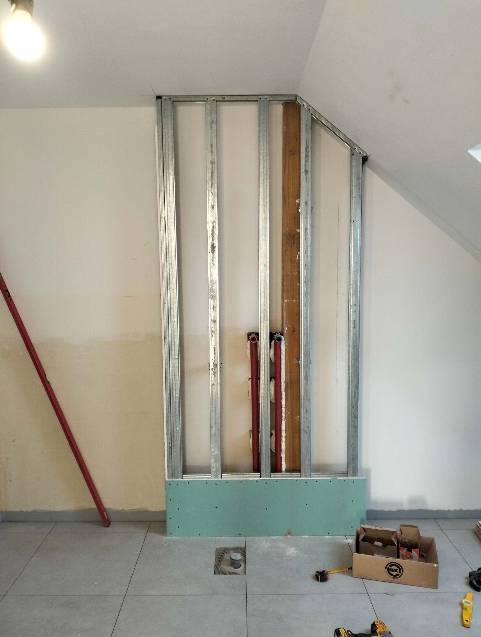 Montaż stelażu z profili stalowych i płyty GK w łazience na poddaszu. Widoczne rury instalacji wodnej. Częściowo położone płytki podłogowe, narzędzia pracy.