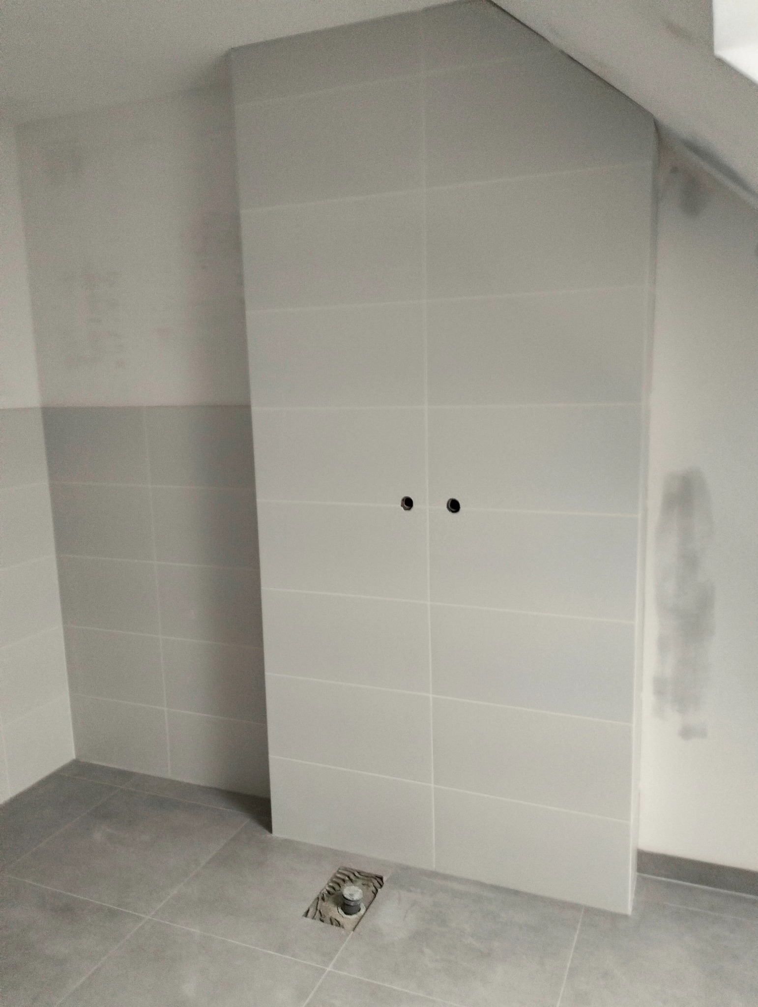 Jasne, prostokątne płytki ścienne i podłogowe w łazience, widoczne otwory na armaturę i odpływ podłogowy. Minimalistyczny design z szarymi fugami.