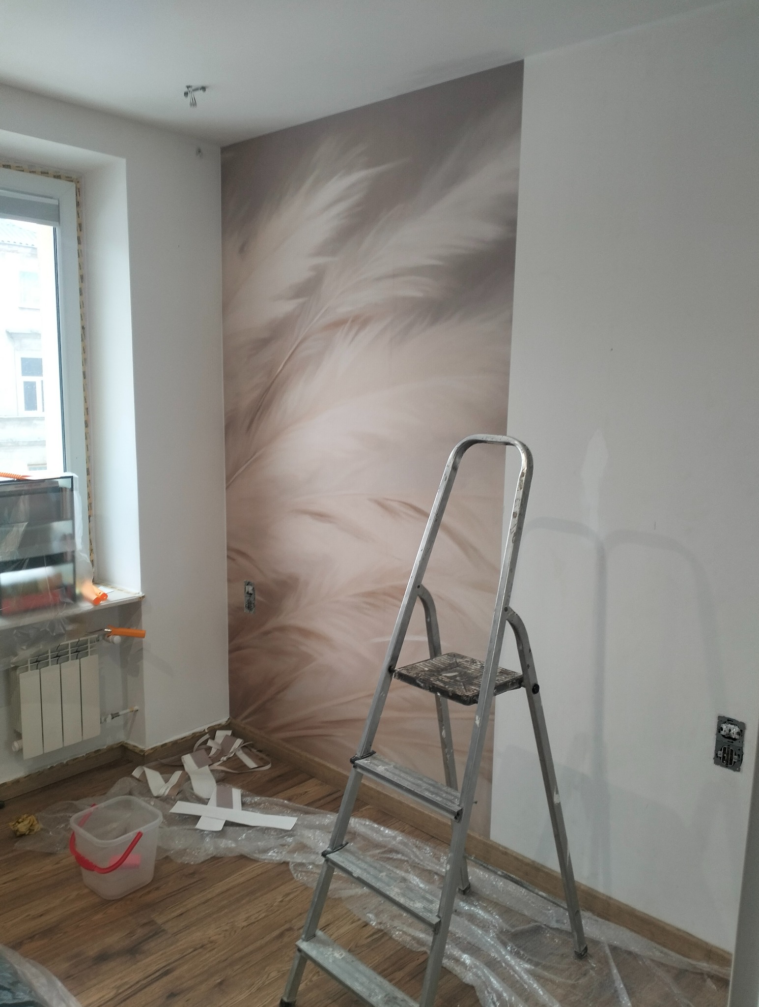 Pokój po tapetowaniu z motywem piór, widoczna drabina, resztki tapety na podłodze. Ujęcie z bliska, ukazujące detale wykończenia i etap prac.