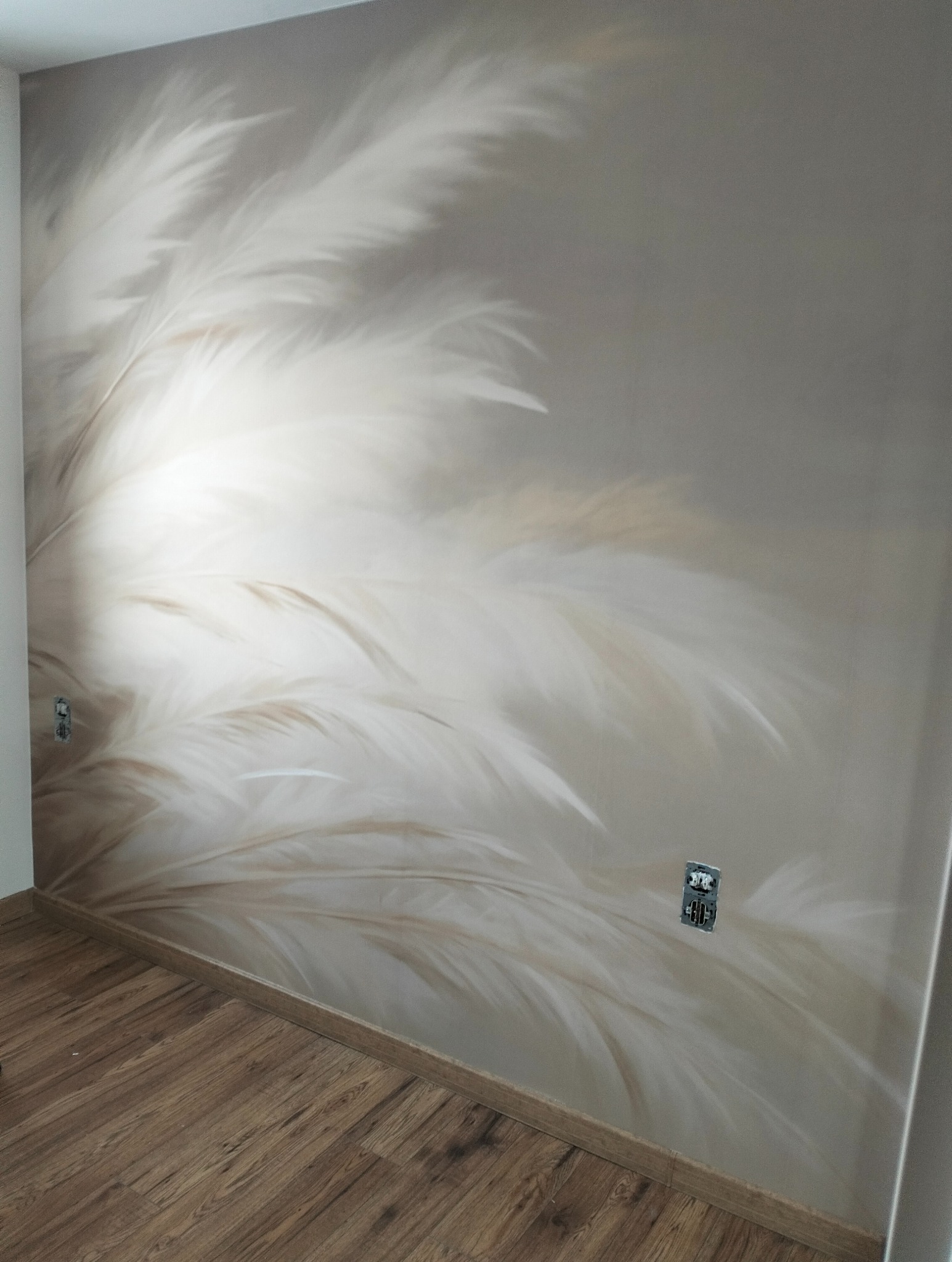 Ściana w pokoju po malowaniu z motywem piór, widoczne gniazdka elektryczne i panele podłogowe. Jasne, stonowane kolory.