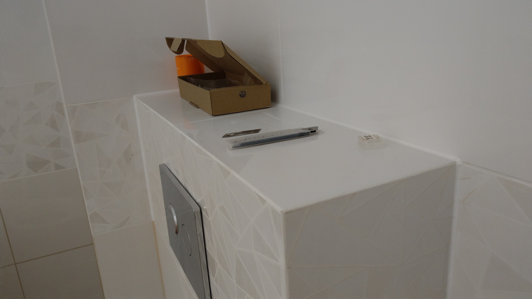 Biała obudowa stelaża WC z geometrycznym wzorem płytek, na której leżą nożyki segmentowe, otwarte pudełko kartonowe i mała kostka, ściany wykończone białymi płytkami.