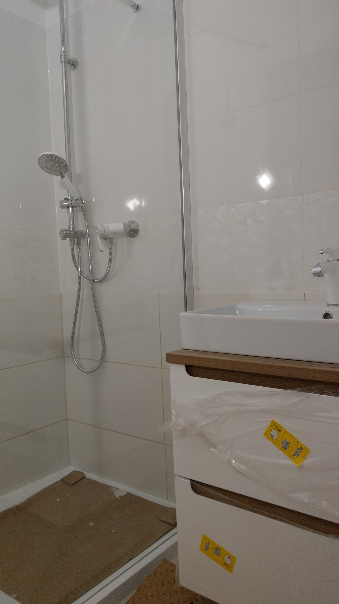 Umywalka z szafką w folii ochronnej, kabina prysznicowa z chromowaną baterią, białe płytki na ścianach z delikatnym wzorem geometrycznym, karton na podłodze w trakcie remontu łazienki.