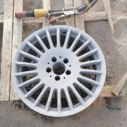 Aluminiowa felga samochodowa po obróbce strumieniowo-ściernej, leżąca na drewnianej palecie obok pistoletu do piaskowania.