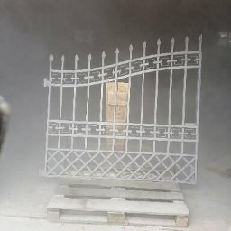 Aleksandra Kowalczyk - Gritter - Metalowa brama ogrodowa z dekoracyjnymi elementami, umieszczona na palecie i otoczona mgłą, prawdopodobnie podczas procesu nakładania powłoki ochronnej.