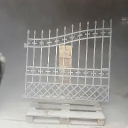 Metalowa brama ogrodowa z dekoracyjnymi elementami, umieszczona na palecie i otoczona mgłą, prawdopodobnie podczas procesu nakładania powłoki ochronnej.