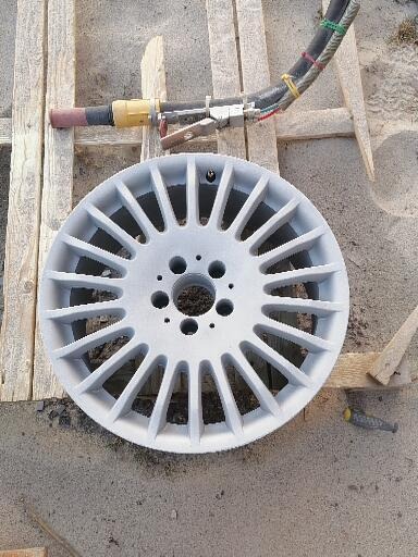 Aluminiowa felga samochodowa po obróbce strumieniowo-ściernej, leżąca na drewnianej palecie obok pistoletu do piaskowania.