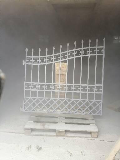 Metalowa brama ogrodowa z dekoracyjnymi elementami, umieszczona na palecie i otoczona mgłą, prawdopodobnie podczas procesu nakładania powłoki ochronnej.