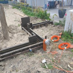 AAP Budownictwo Volodymyr Keryk - Szczegółowy widok szalunków pod fundamenty z widocznym zbrojeniem, pomarańczowymi kablami i rurami kanalizacyjnymi na placu budowy przed domem jednorodzinnym.