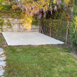 AAP Budownictwo Volodymyr Keryk - Wylana płyta betonowa pod budowę, widok z poziomu trawnika, w tle kwitnąca glicynia na murze i siatce ogrodzeniowej.