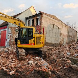 Keryk - Żółta koparka Komatsu na gruzowisku ceglanym, w tle budynek w trakcie rozbiórki, częściowo zburzony, z widocznymi czerwonymi drzwiami remizy strażackiej.