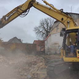 AAP Budownictwo Volodymyr Keryk - Żółta koparka Komatsu w trakcie rozbiórki ceglanego budynku, unosząca gruz. Operator w kabinie, w tle zabudowa miejska i pochmurne niebo. Widoczny pył i resztki materiałów budowlanych.