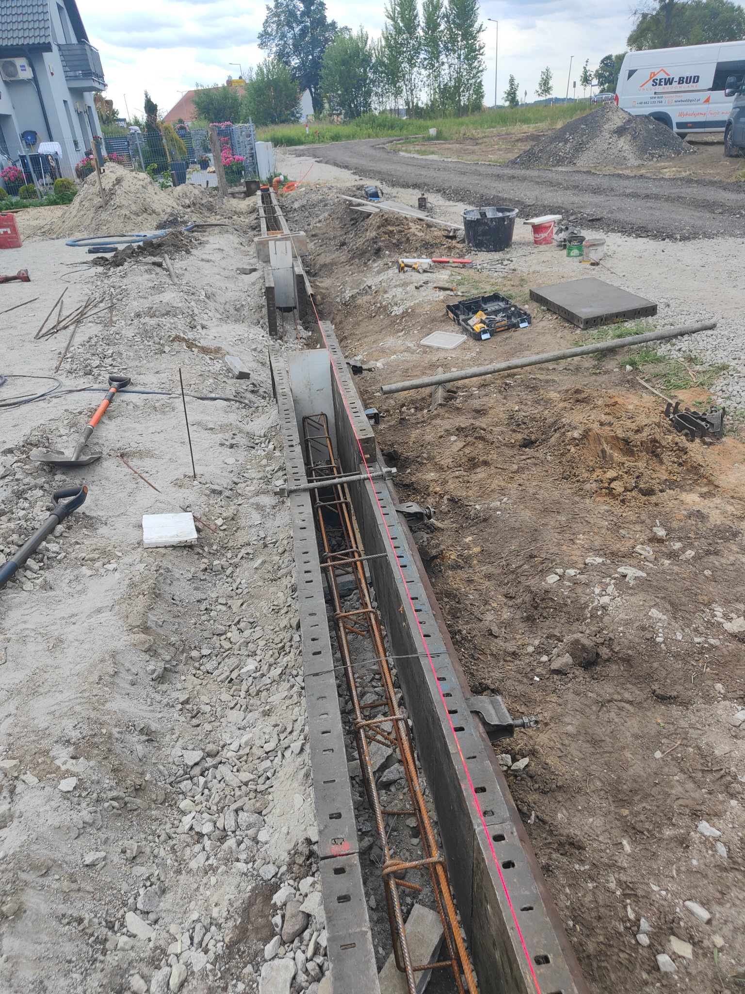 Przygotowanie szalunku i zbrojenia pod wylewkę fundamentu, widoczny laserowy niwelator wyznaczający poziom, narzędzia budowlane w tle, teren budowy.