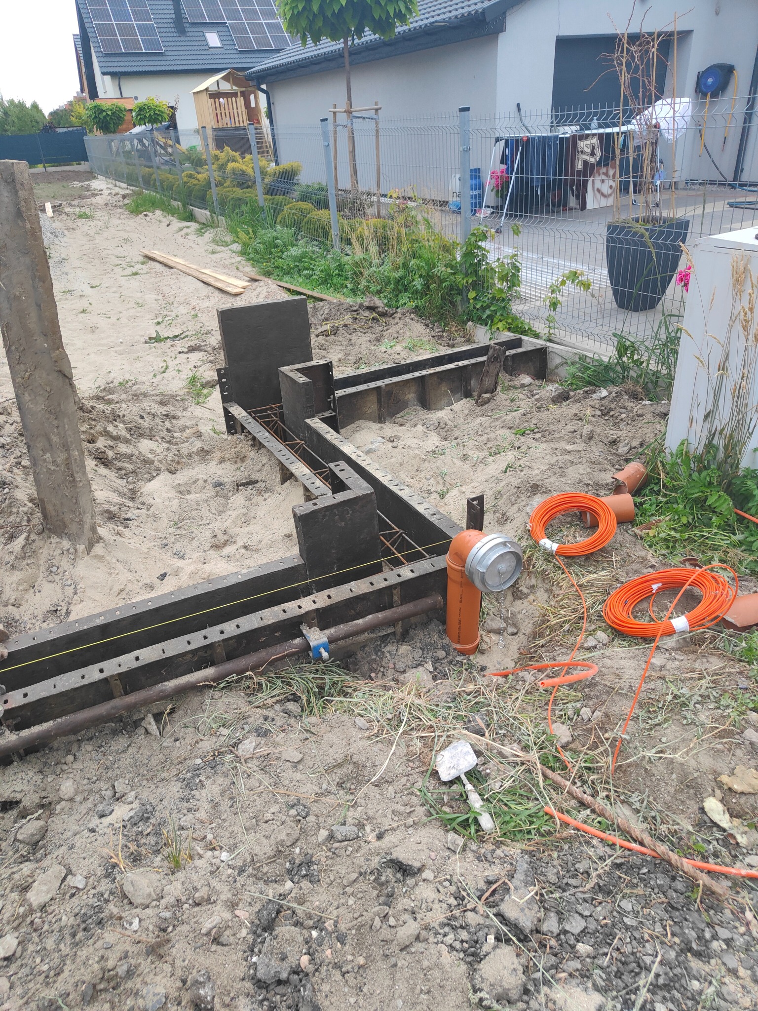 Szczegółowy widok szalunków pod fundamenty z widocznym zbrojeniem, pomarańczowymi kablami i rurami kanalizacyjnymi na placu budowy przed domem jednorodzinnym.