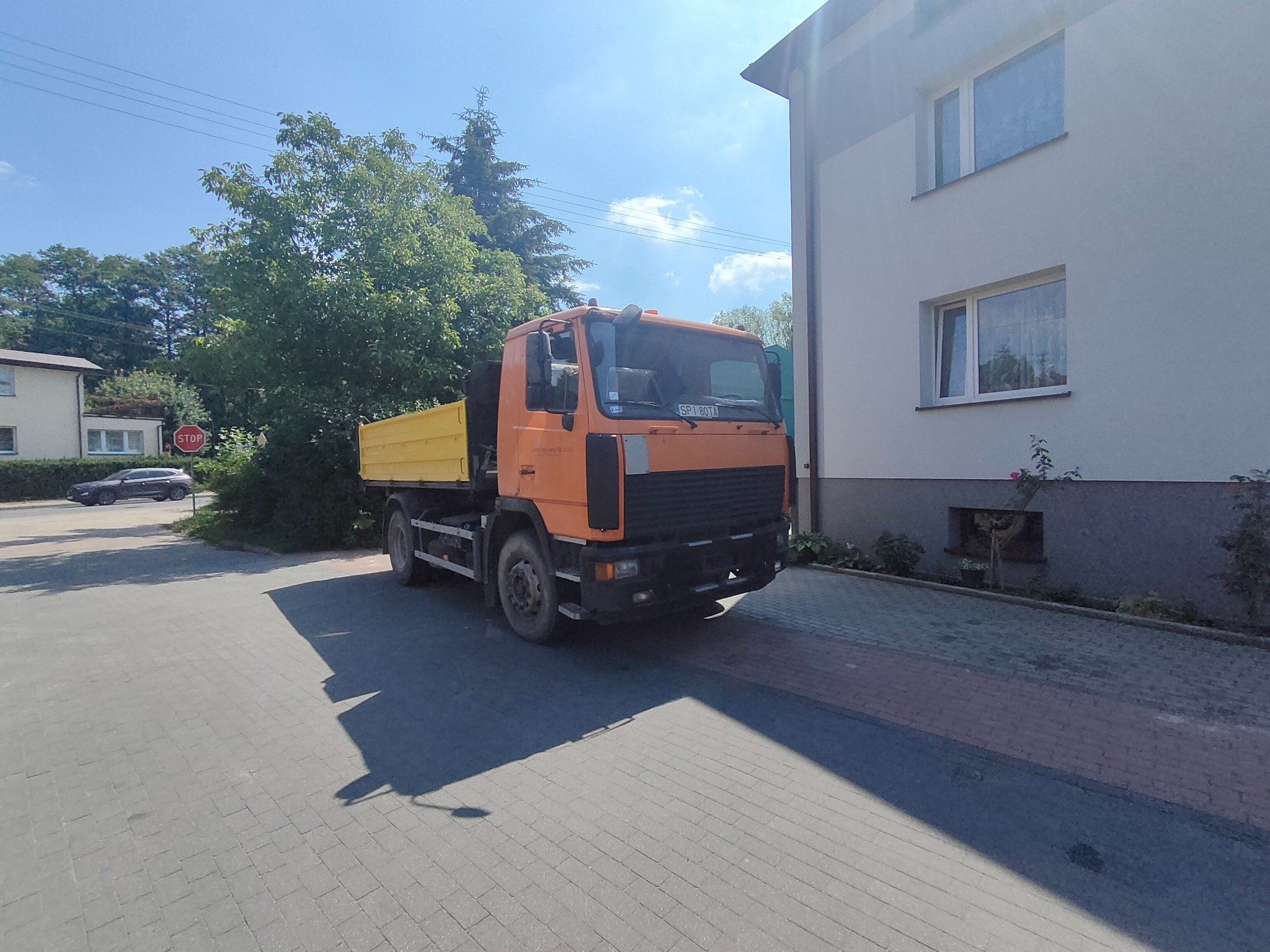 Pomarańczowa wywrotka z żółtą skrzynią ładunkową zaparkowana na kostce brukowej przed budynkiem mieszkalnym w słoneczny dzień.