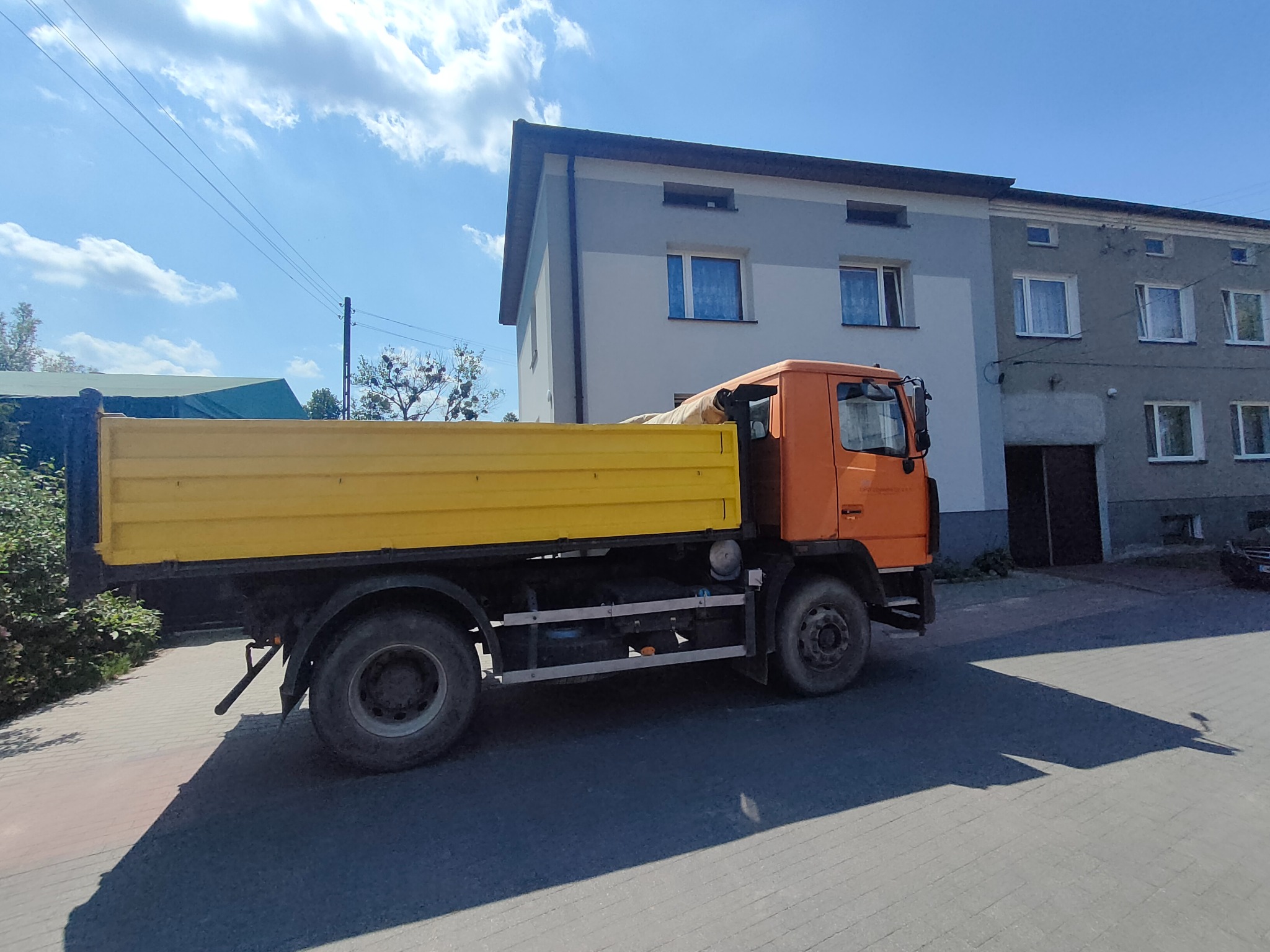 Pomarańczowo-żółta wywrotka zaparkowana na ulicy przed dwupiętrowym budynkiem w słoneczny dzień.