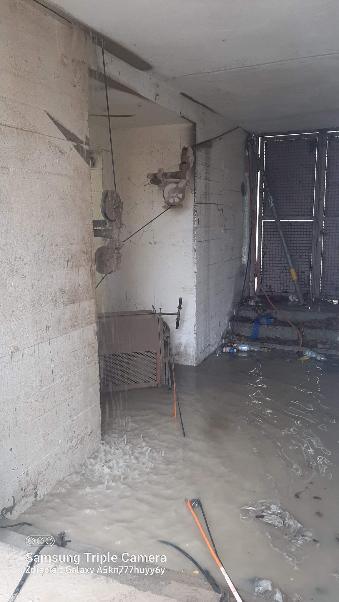 Maszyna do cięcia betonu liną diamentową w trakcie pracy, woda chłodzi linę, widoczne ślady cięcia na ścianie i woda na podłodze.