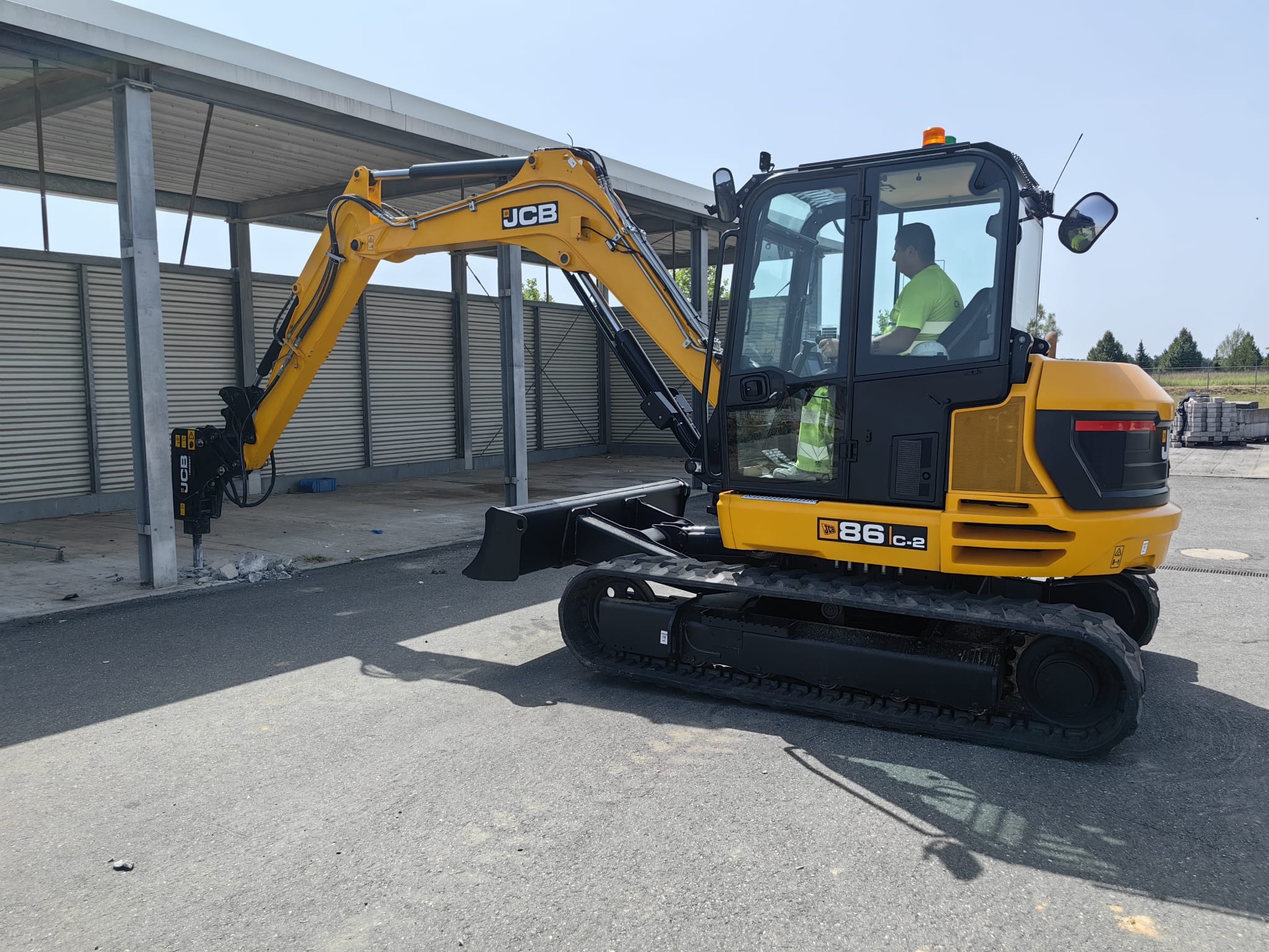 Żółta minikoparka JCB 86C-2 z operatorem w kabinie, wyposażona w młot hydrauliczny, rozbija betonową nawierzchnię pod zadaszeniem.