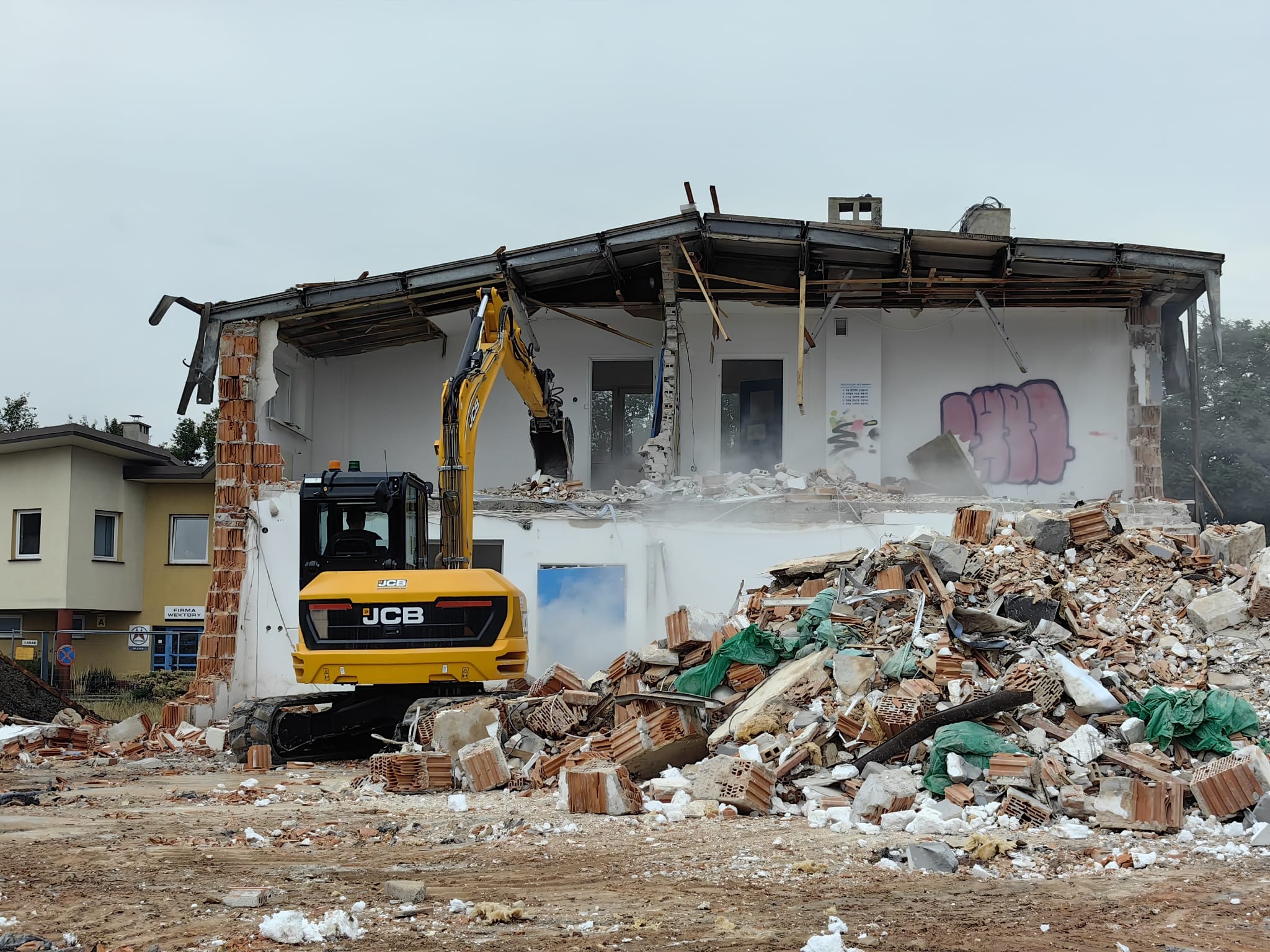 Żółta koparka JCB podczas rozbiórki budynku z białymi ścianami, widoczne gruzy i elementy konstrukcyjne, graffiti na ścianie.