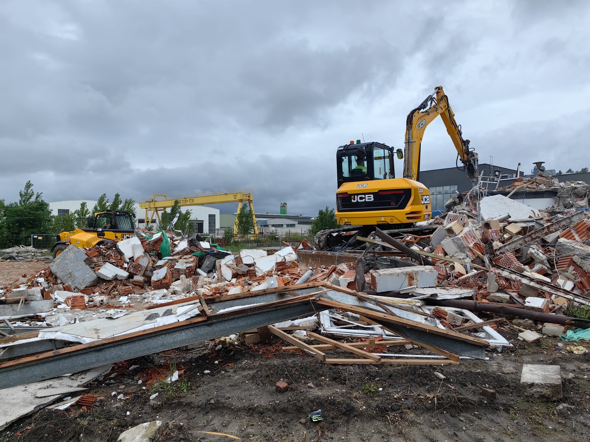 Żółta koparka JCB na tle gruzowiska po rozbiórce budynku, z widocznym fragmentem stalowej konstrukcji i innym sprzętem budowlanym w tle, pochmurne niebo.