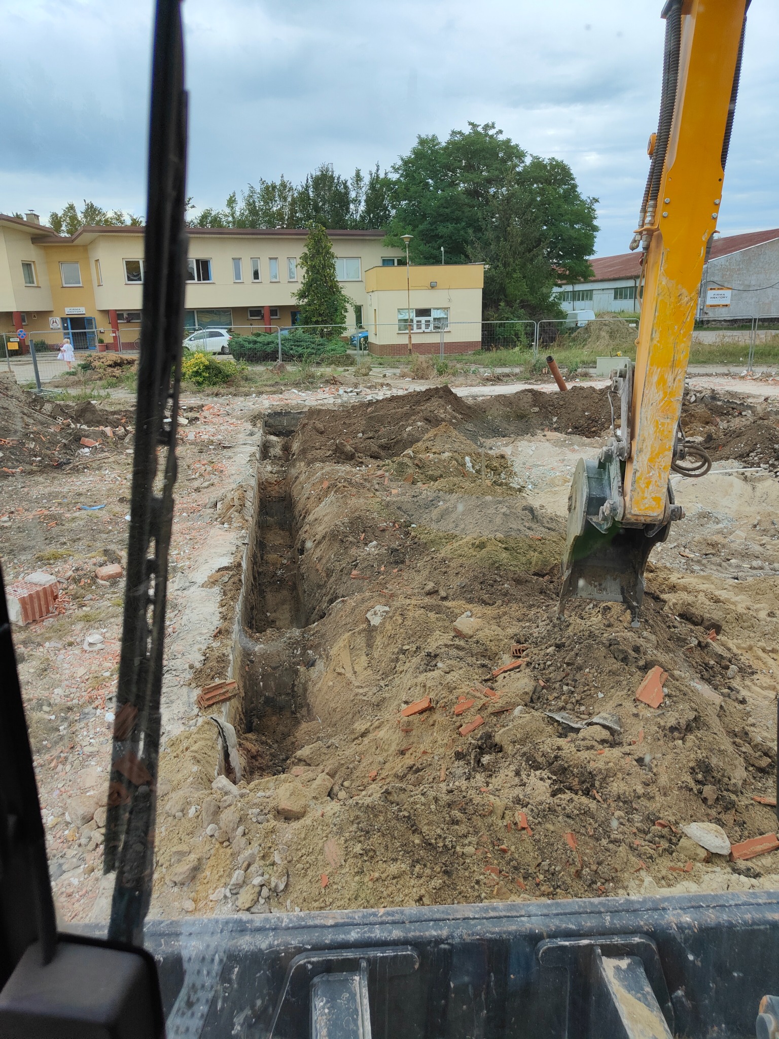 Wykopany wąski rów w ziemi z widocznymi fragmentami cegieł, wykonany koparką, widok z kabiny operatora na tle zabudowań.