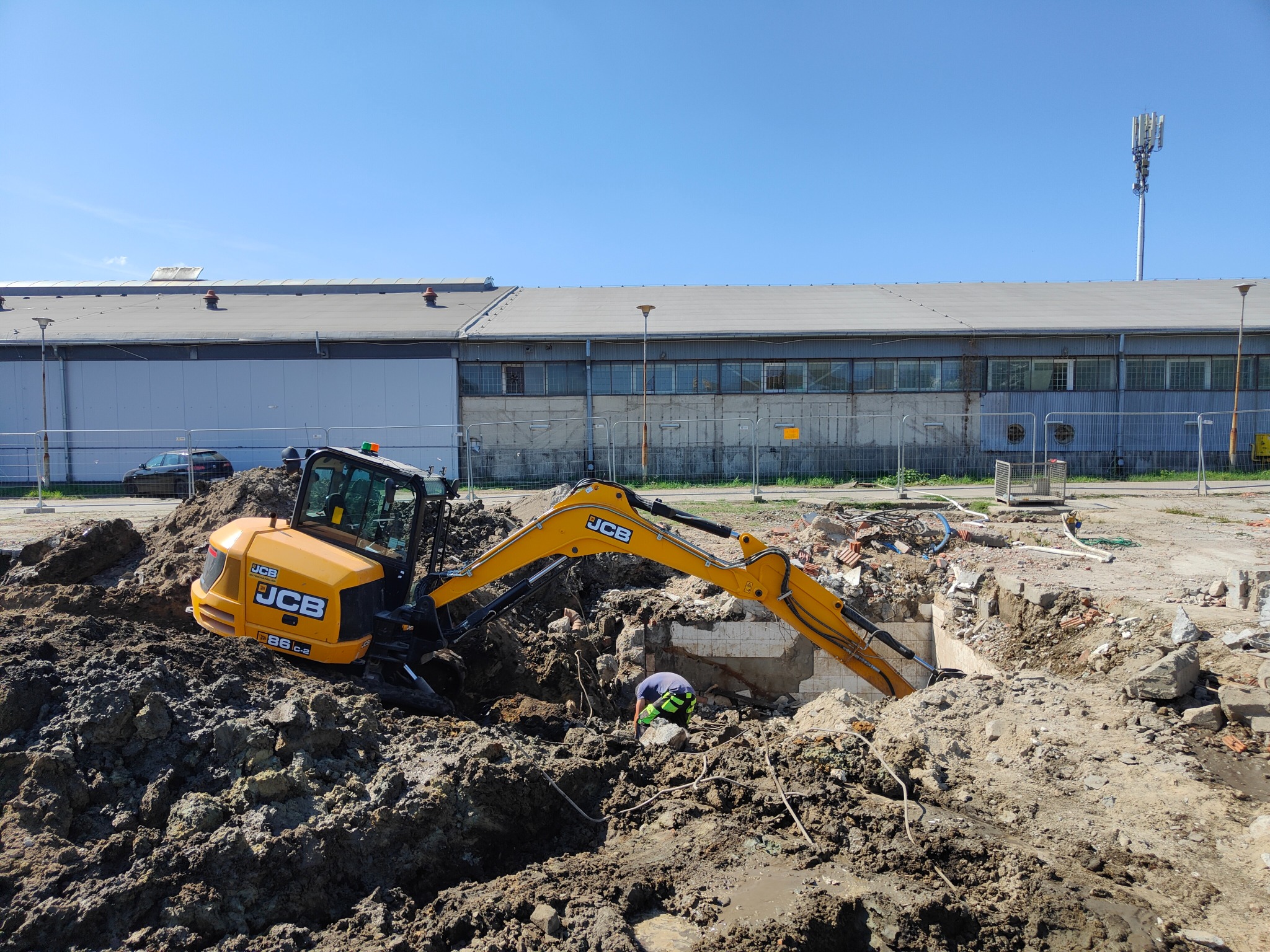 Żółta koparka JCB 86C-2 pracuje w wykopie, widoczny fragment zburzonej ściany, pracownik w kasku i odblaskowej kamizelce w dole, w tle budynek przemysłowy i maszt telekomunikacyjny.
