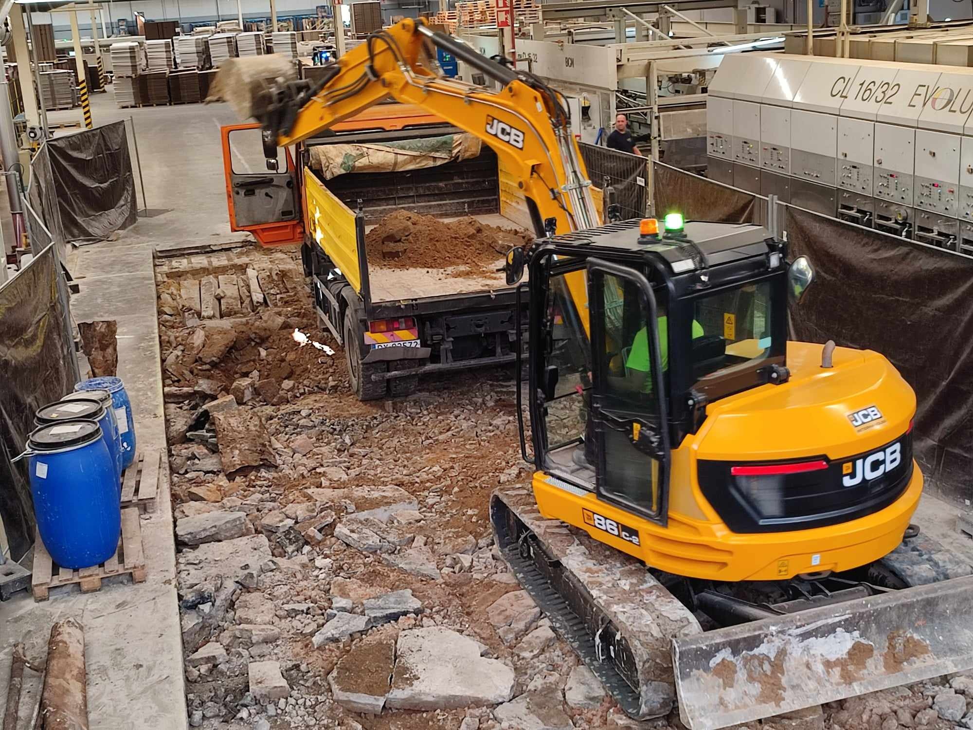 Żółta koparka JCB ładuje urobek do pomarańczowej ciężarówki wewnątrz hali produkcyjnej; w tle widoczne niebieskie beczki i maszyny przemysłowe.