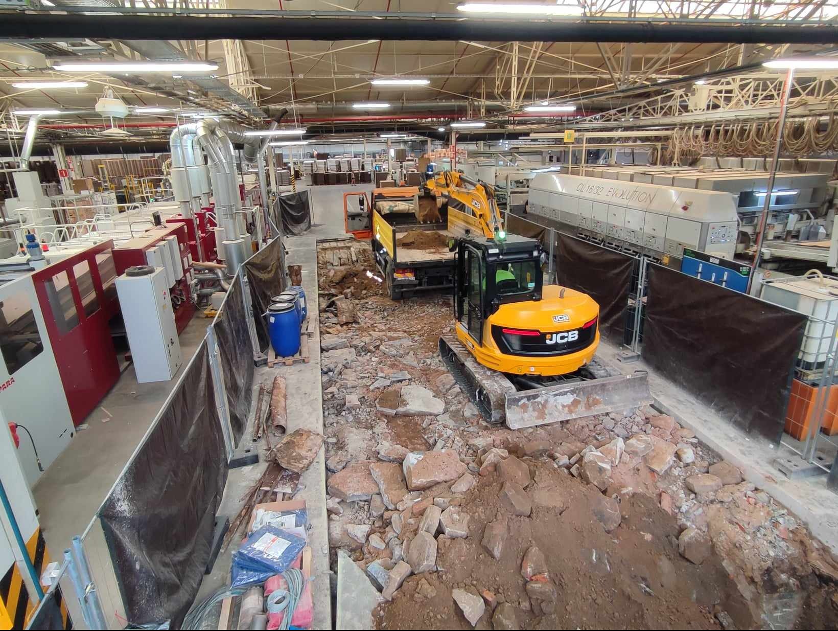 Żółta koparka JCB i wywrotka podczas prac ziemnych wewnątrz hali przemysłowej, gruz i ziemia w wykopie, widoczne elementy maszyn i instalacji.