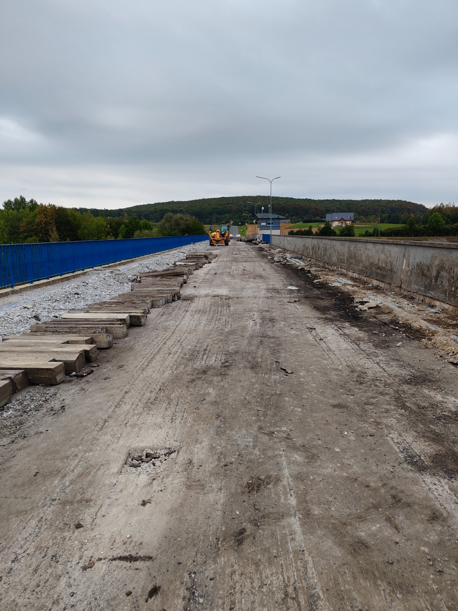 Rozbiórka mostu w toku: zerwana nawierzchnia, gruz, fragmenty betonowe, w tle koparka i zieleń. Widok z perspektywy jezdni.