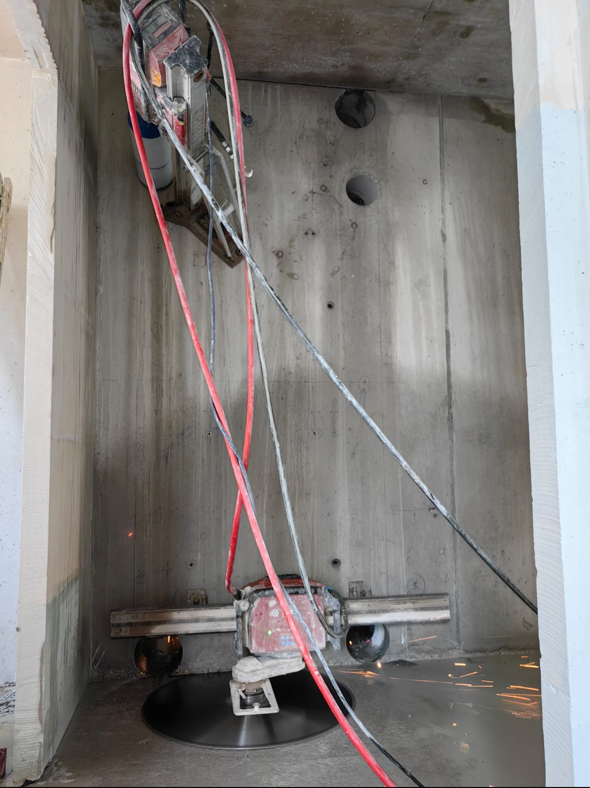 Cięcie betonu piłą diamentową w pomieszczeniu, widoczne iskry. Na ścianie ślady po wierceniu. Czerwone i szare przewody. Betonowa konstrukcja.