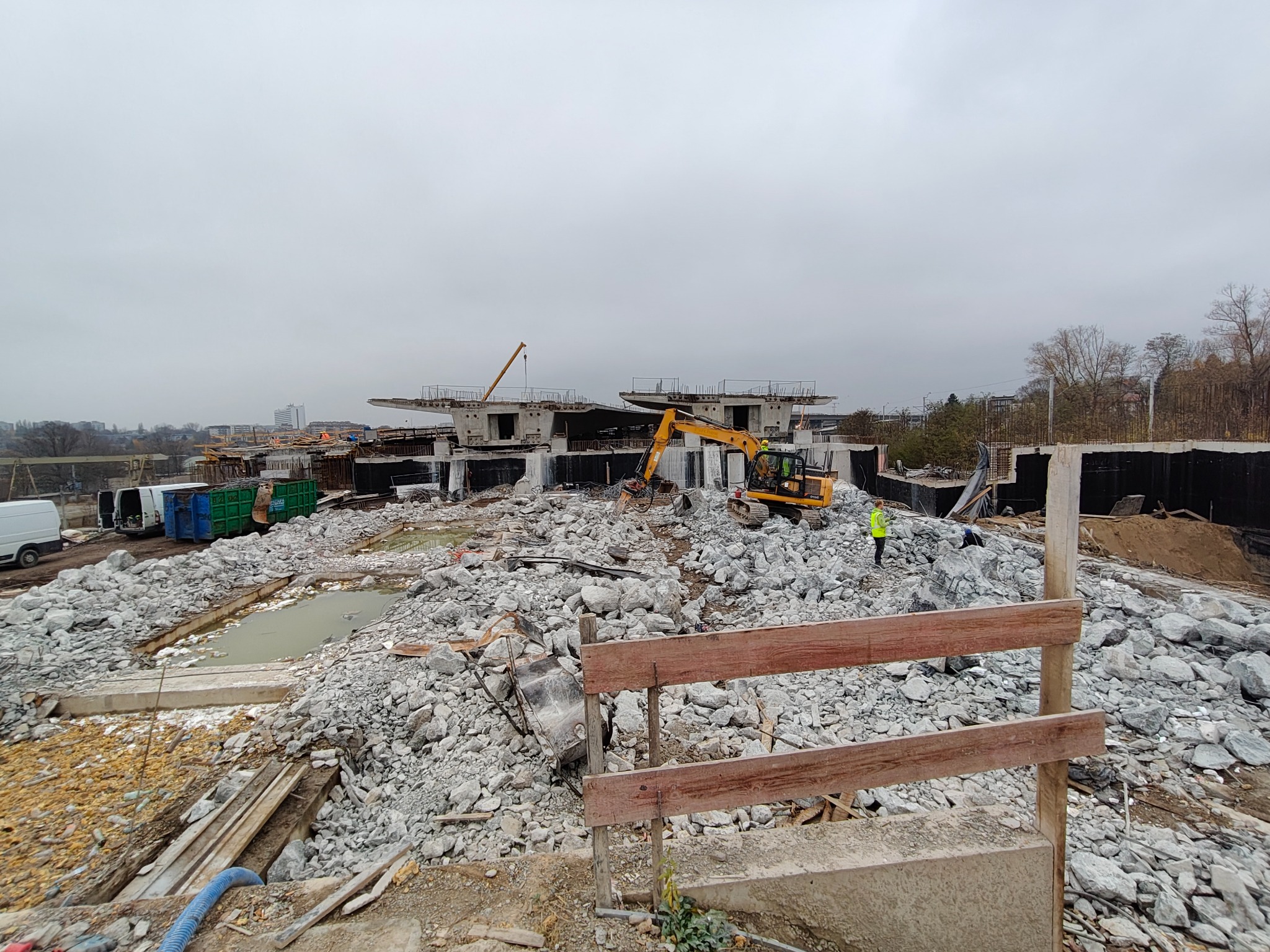 Żółta koparka na placu budowy z gruzem, w tle betonowe konstrukcje i zabudowa miejska pod pochmurnym niebem. Widoczne elementy zabezpieczeń i pracownicy w odblaskowych kamizelkach.