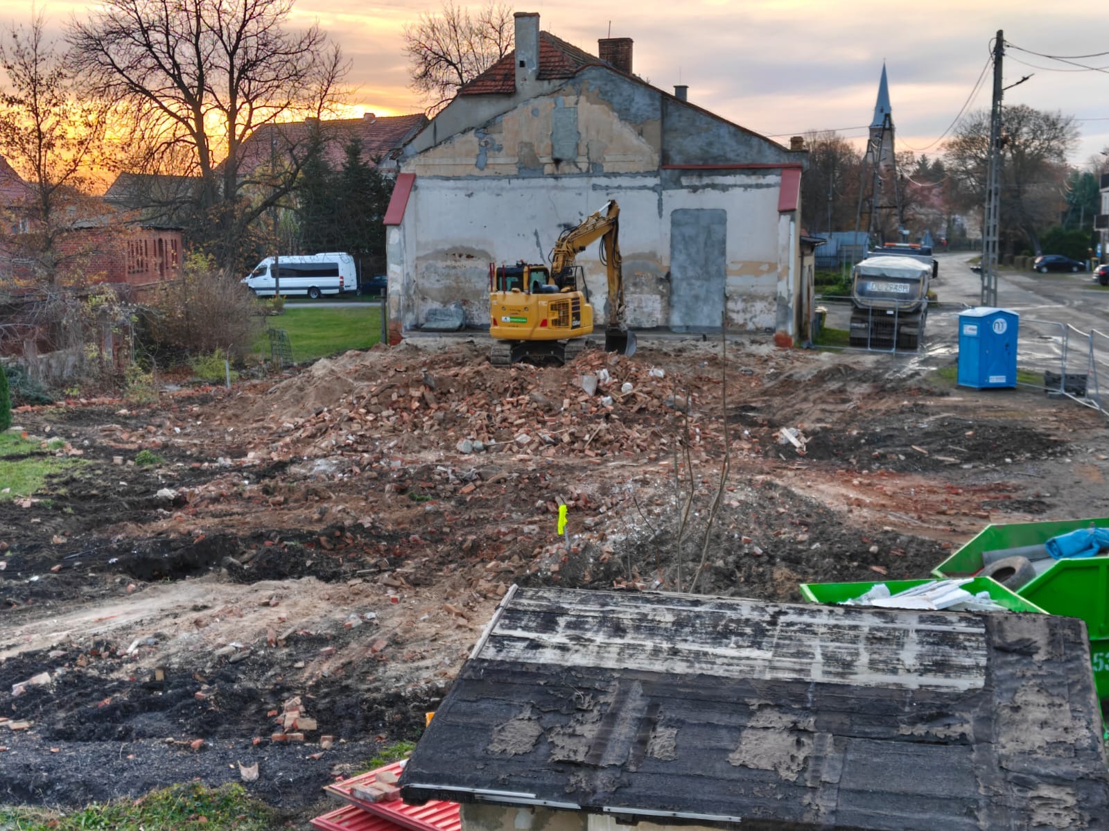 Żółta koparka na placu budowy po rozbiórce, gruz i ziemia, w tle budynek z odsłoniętą ścianą i kościół. Widok z góry, pochmurne niebo.