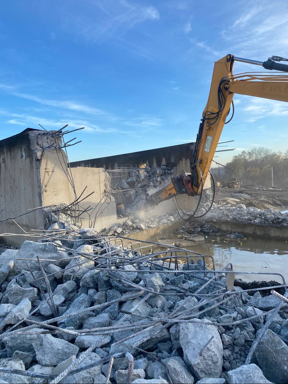 Żółty koparko-ładowarka z młotem pneumatycznym rozbija betonową konstrukcję mostu. W tle błękitne niebo i drzewa. Gruz i woda w dolnej części kadru.