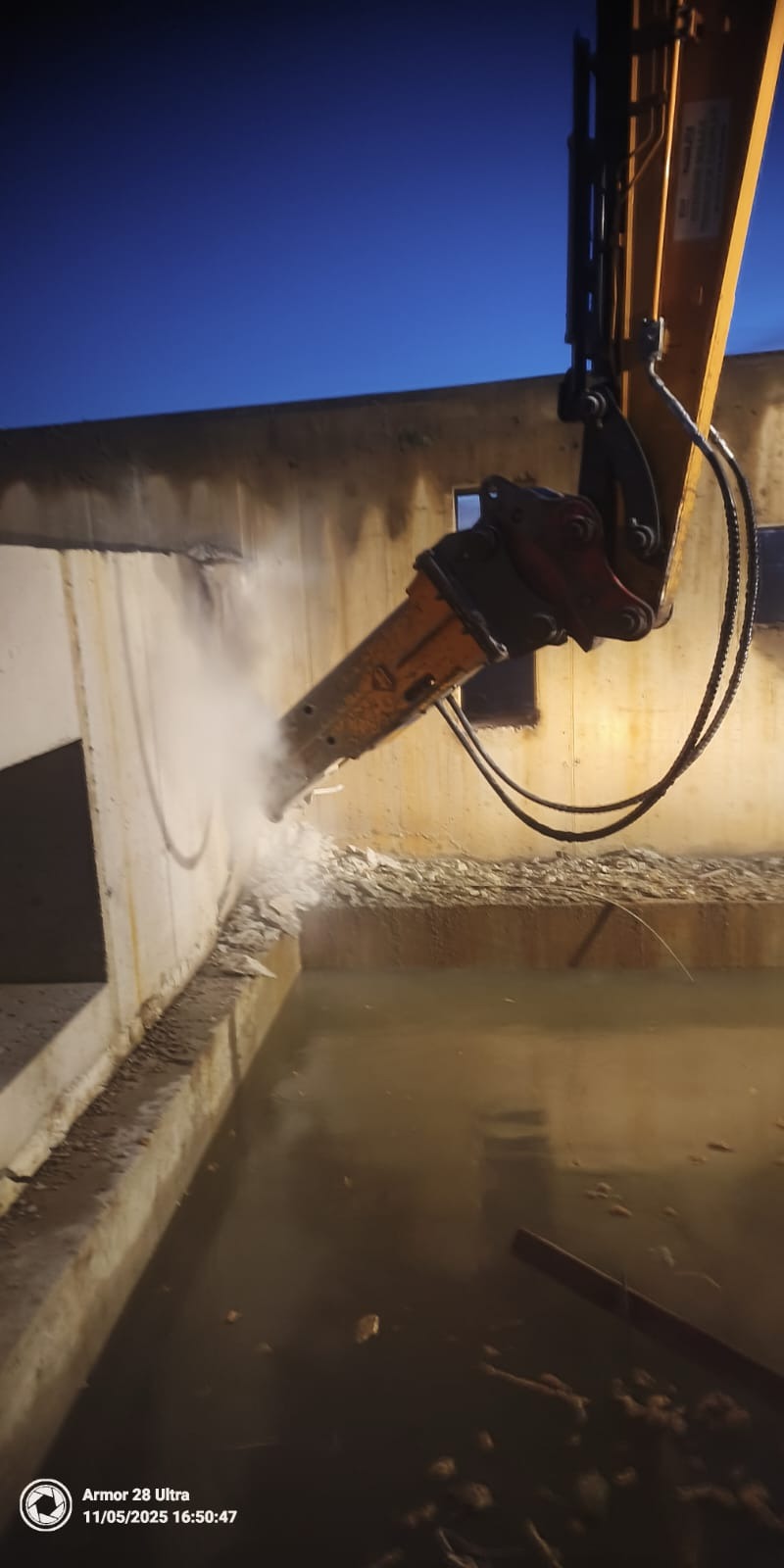 Hydrauliczny młot wyburzeniowy kruszy betonową ścianę. Wokół unosi się pył, a w dole widać wodę. Praca w toku, o zmierzchu.