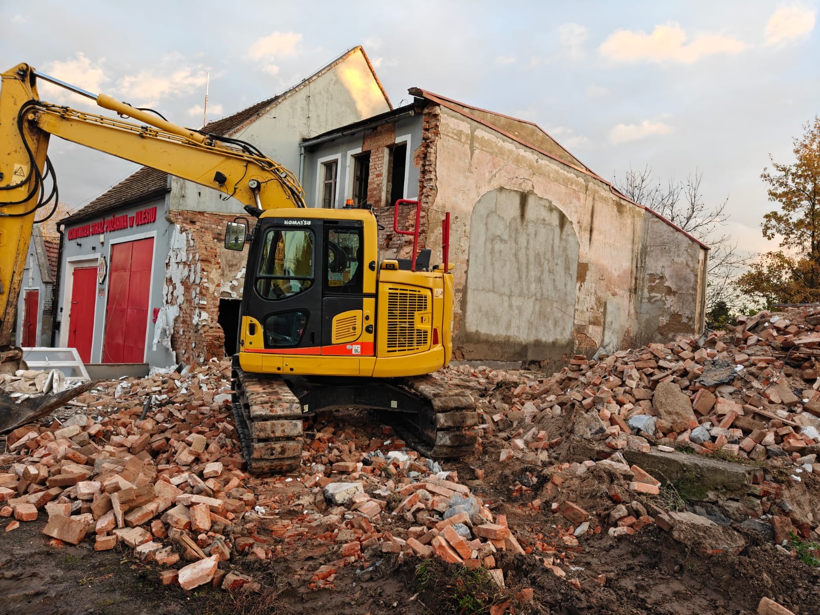 Żółta koparka Komatsu na gruzowisku ceglanym, w tle budynek w trakcie rozbiórki, częściowo zburzony, z widocznymi czerwonymi drzwiami remizy strażackiej.