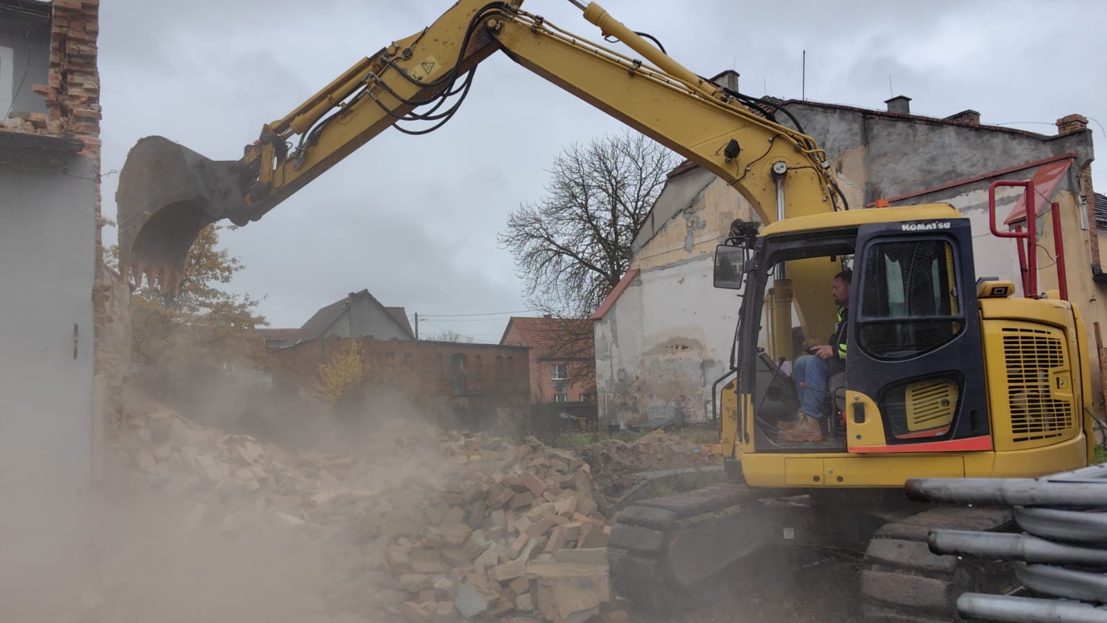Żółta koparka Komatsu w trakcie rozbiórki ceglanego budynku, unosząca gruz. Operator w kabinie, w tle zabudowa miejska i pochmurne niebo. Widoczny pył i resztki materiałów budowlanych.