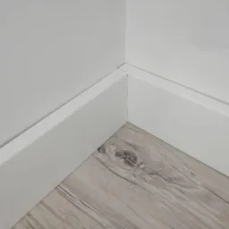 Połączenie ściany i podłogi z szarych paneli imitujących drewno, wykończone białą listwą przypodłogową w rogu pomieszczenia.