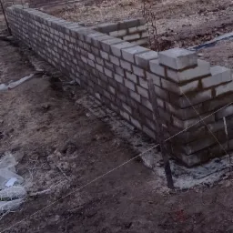 Wznoszenie muru fundamentowego z bloczków betonowych, widoczne sznurki traserskie i drewniane kołki, przygotowanie do dalszych prac budowlanych.