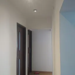 Korytarz z drewnopodobną podłogą, dwiema parami drzwi z przeszkleniami i oświetleniem punktowym w suficie, ściany pomalowane na biało i błękitno.