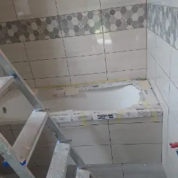 Wnętrze łazienki w trakcie remontu: widoczna drabina aluminiowa, zabezpieczona wanna, częściowo ułożone płytki ścienne z dekoracyjnym pasem, wystające przewody elektryczne i rury wodne.
