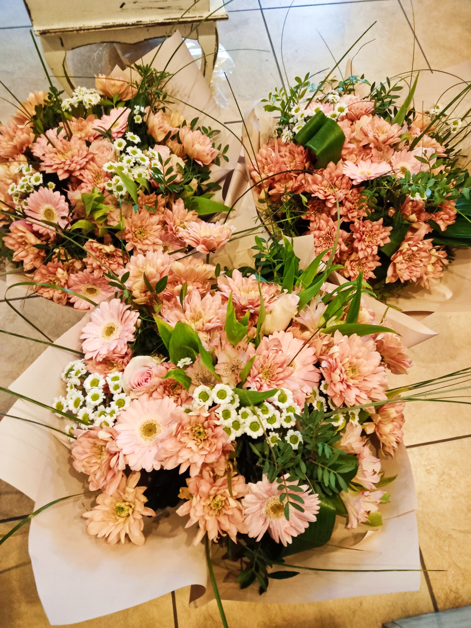 Trzy bukiety z różowych gerber, białych margaretek i zielonych dodatków, owinięte w jasny papier, leżące na kafelkach.