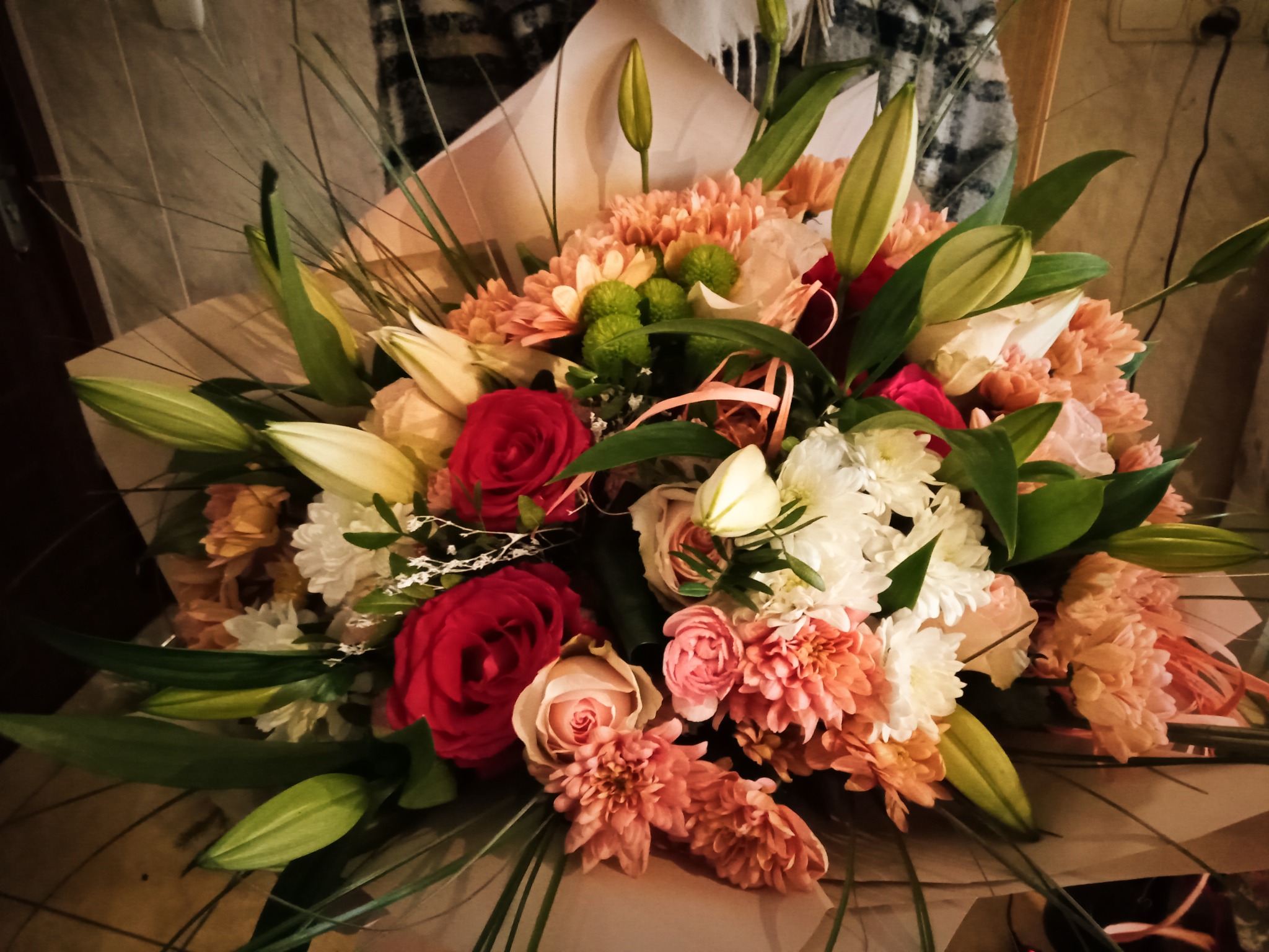 Duży bukiet mieszanych kwiatów w odcieniach czerwieni, bieli, różu i zieleni, z przewagą goździków, róż i lilii, owinięty w jasny papier.