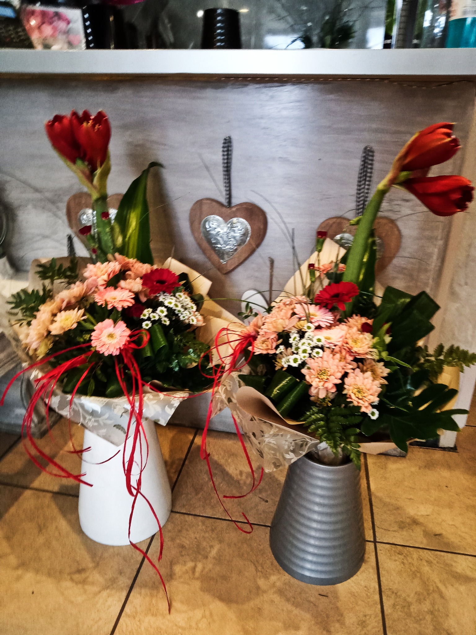 Dwa bukiety kwiatów w ozdobnych wazonach, z amarylisami i gerberami, zapakowane w papier dekoracyjny, ustawione na brązowej podłodze z płytek, tło stanowią drewniane serca na ścianie.