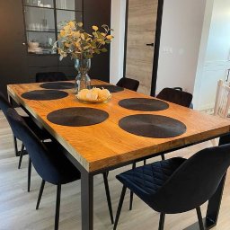 Hardwood Classic - Stół z dębowym blatem i metalowymi nogami, otoczony czarnymi krzesłami z weluru, nakryty czarnymi podkładkami. Na stole wazon z gałązkami i dekoracyjne dynie. W tle zabudowa kuchenna...