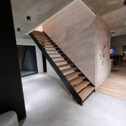 Nowoczesne schody z drewnianymi stopniami i czarną metalową konstrukcją w minimalistycznym wnętrzu z betonowym sufitem i ścianami o surowym wykończeniu.