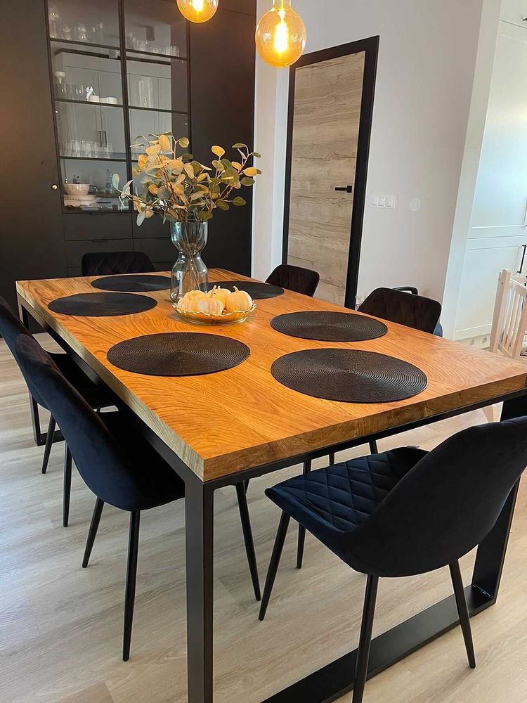 Stół z dębowym blatem i metalowymi nogami, otoczony czarnymi krzesłami z weluru, nakryty czarnymi podkładkami. Na stole wazon z gałązkami i dekoracyjne dynie. W tle zabudowa kuchenna...