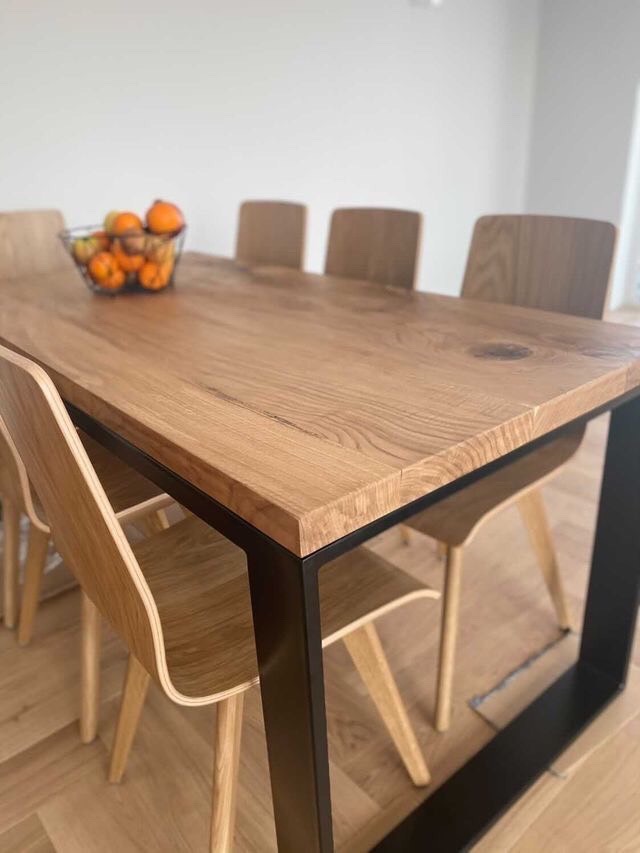 Stół jadalny z blatem z litego drewna o wyraźnym usłojeniu, z czarnymi, metalowymi nogami w kształcie litery U, otoczony drewnianymi krzesłami, na blacie miska z owocami.