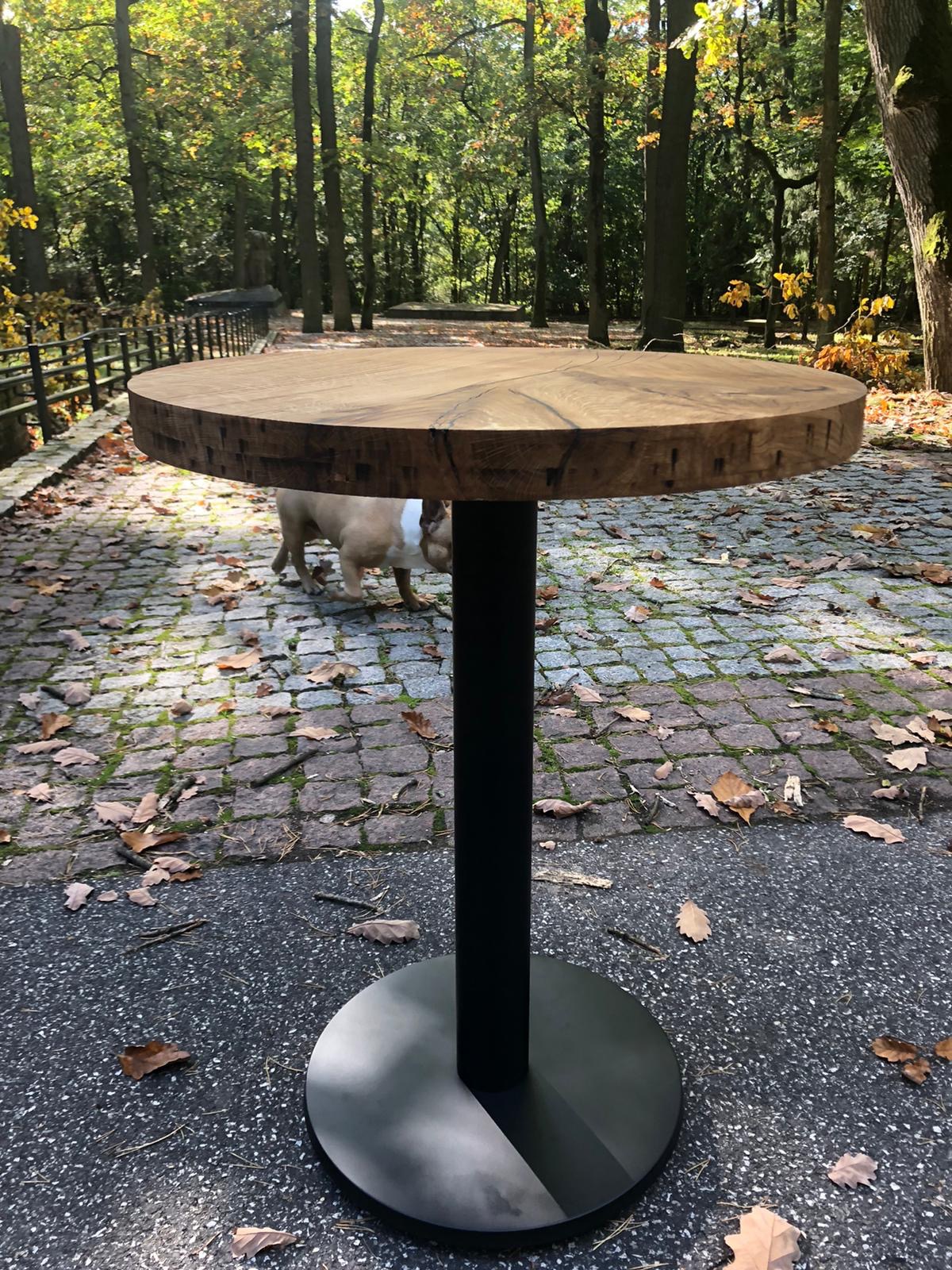 Okrągły stół barowy z blatem z litego drewna i czarną, metalową nogą, ustawiony na zewnątrz na tle jesiennego parku z psem w tle.
