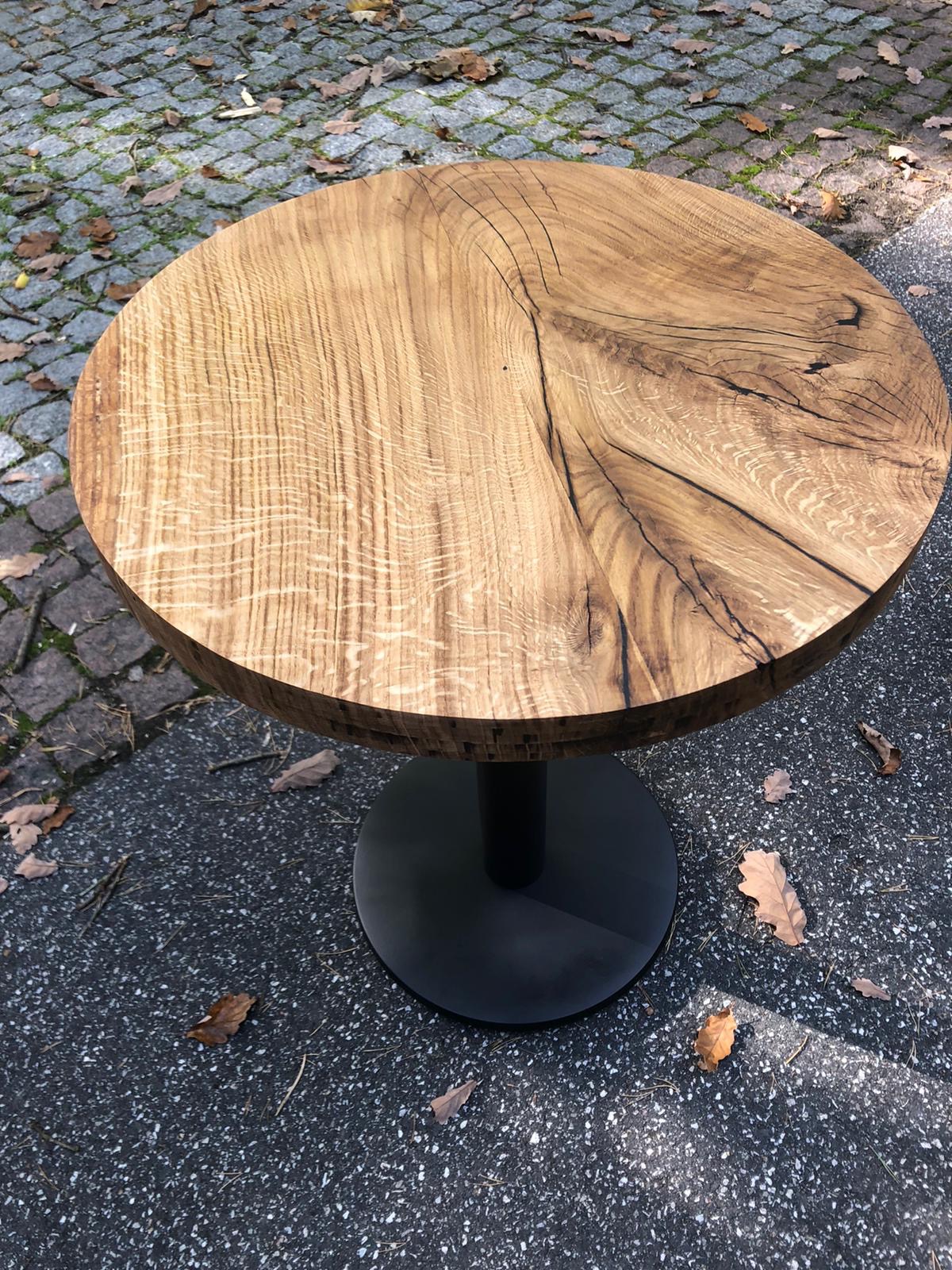 Okrągły stolik kawowy z blatem z litego drewna o wyraźnym usłojeniu i czarną, metalową podstawą, ustawiony na zewnątrz na tle kostki brukowej i opadłych liści.